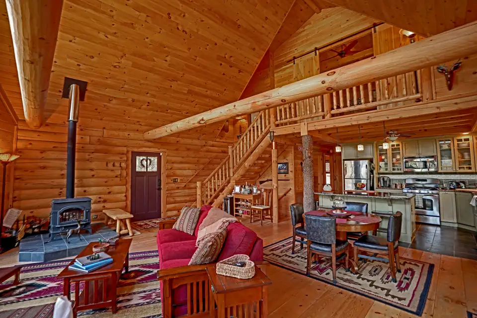 Log cabin great room with wood burning stove, log staircase, loft, and kitchen beyond